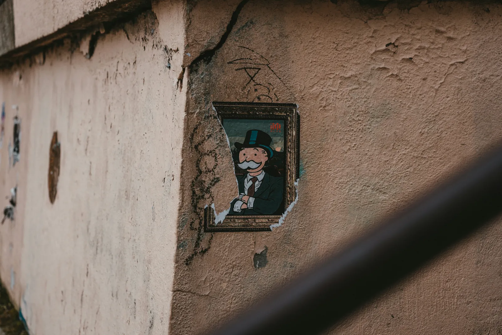 A high-zoom photo of what appears to be an exterior wall; There is graffiti, and the wall has cracks in it and the finish itself. There is a fake framed portrait of the monopoly guy pasted onto it; It is partially torn off.